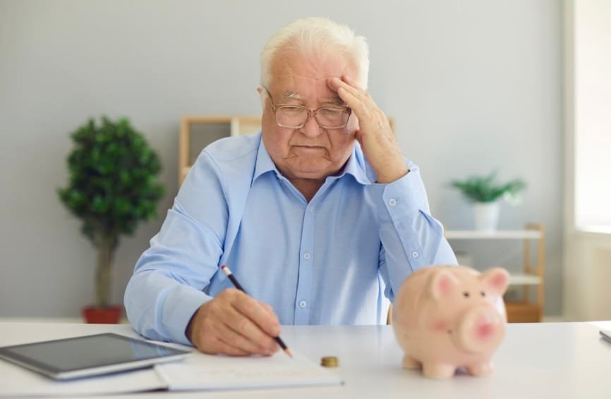 Homme senior inquiet calculant son budget retraite devant une tirelire cochon